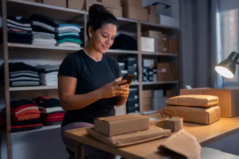 Dona de loja virtual confere o celular no estoque, entre caixas de pedidos