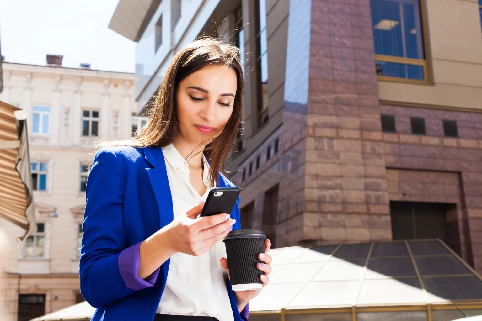 Mulher de blazer azul confere o celular na rua, com um copo de café na outra mão