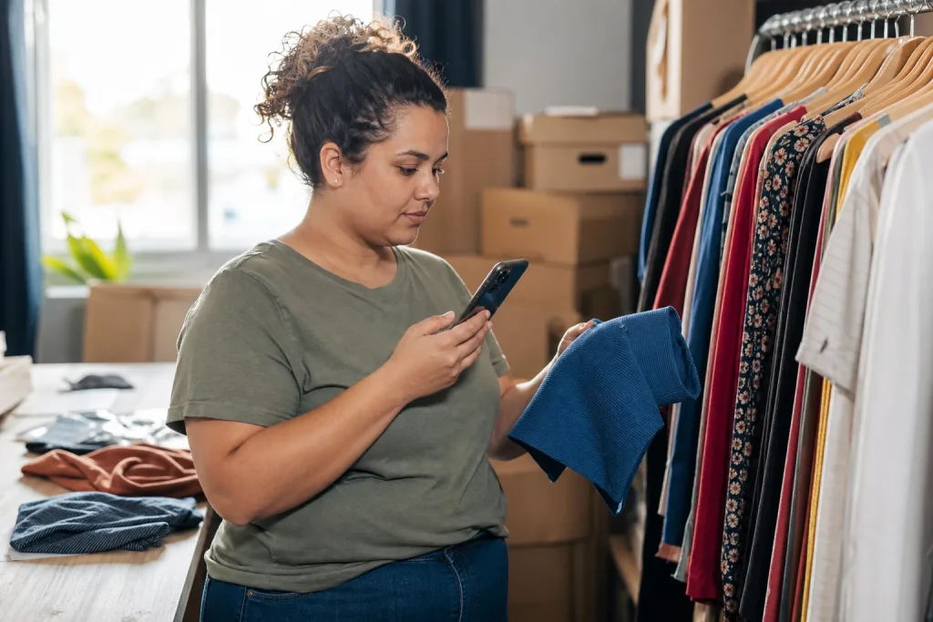 Lojista brasileira conferindo produtos no celular dentro de um pequeno estoque de roupas