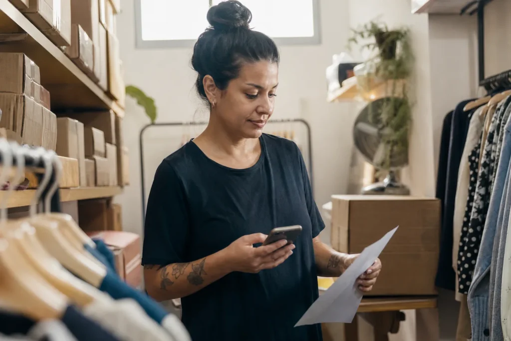 Lojista brasileira confere o celular em um estoque de roupas enquanto segura uma lista de pedidos