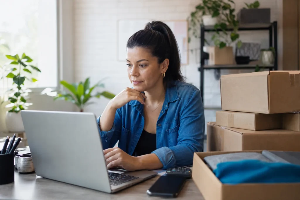 Lojista brasileira analisa painel de anúncios no notebook ao lado de caixas de produtos