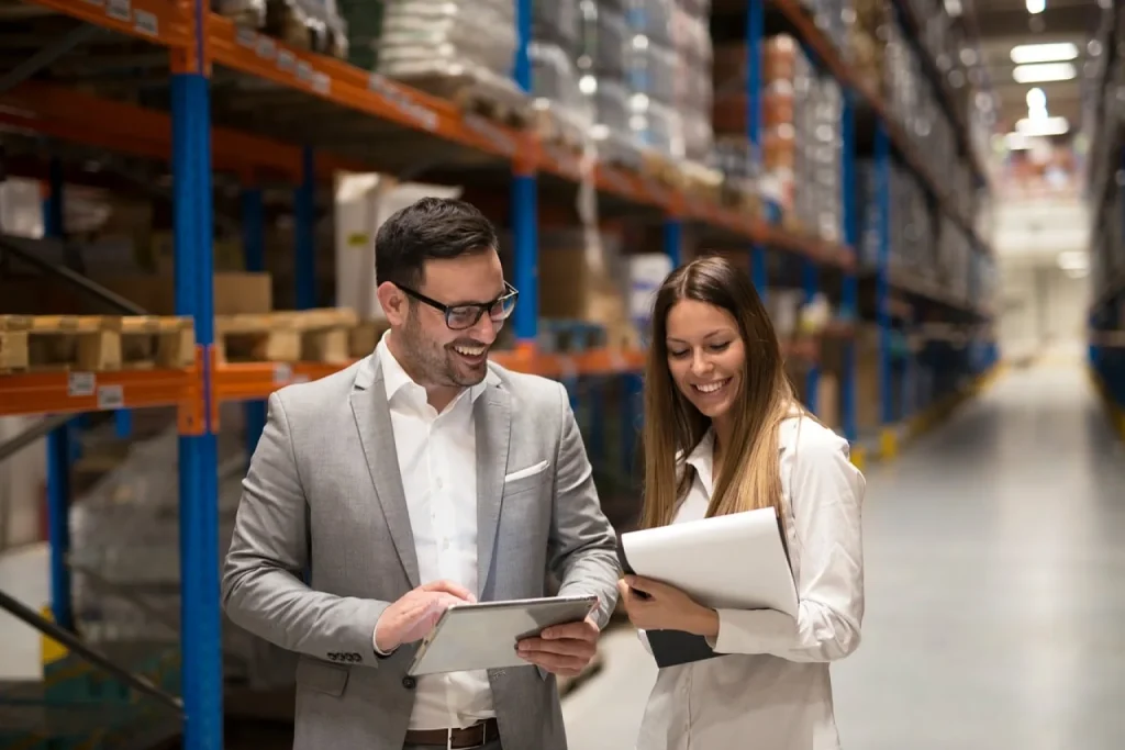 Dona de loja virtual confere planilha de códigos de produtos com um tablet no estoque, preparando a migração de plataforma